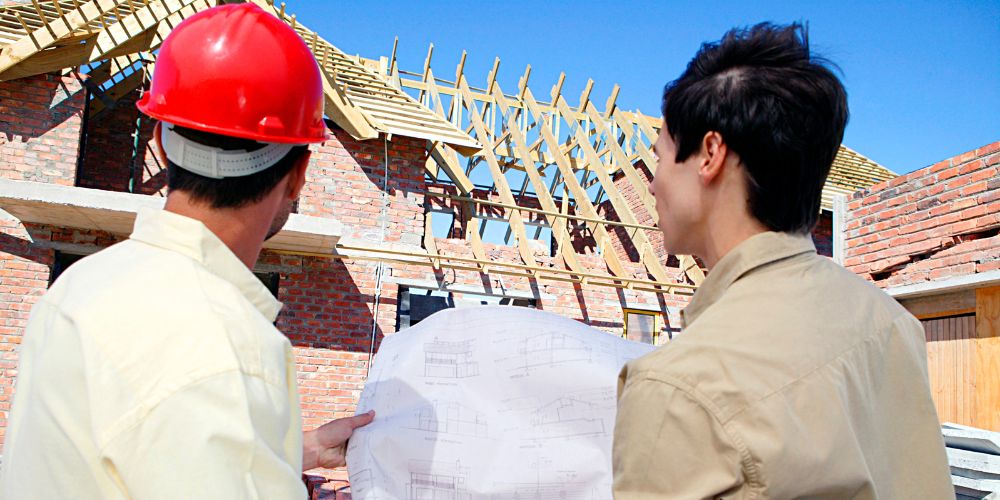 Man holding a roofing plan with a client, facing a house with a roof under construction